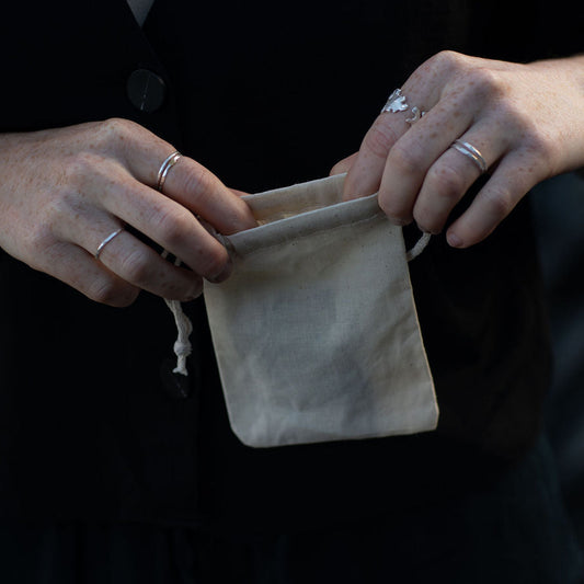 A close-up of a person wearing a black cardigan and holding a small beige fabric drawstring pouch. The person is also wearing silver rings and a gold necklace.