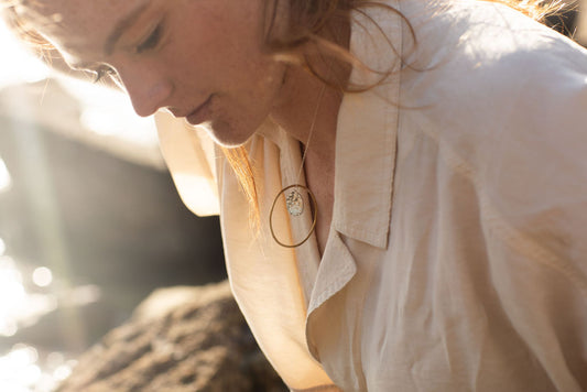 Woman wearing a necklace with a pendant, standing outdoors with sunlight.