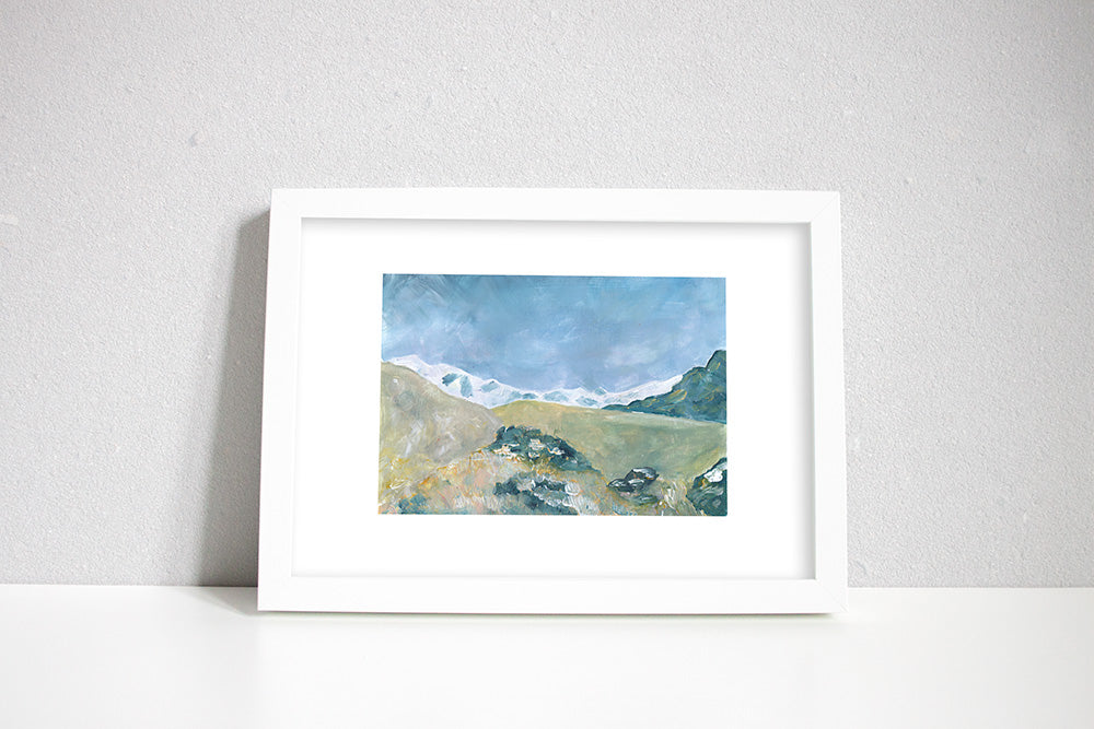 A framed painted picture of the Himalayan mountains, with a village in the foreground and white snow capped mountains in background. 
