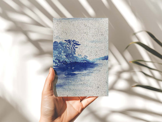 Hand holding a portrait painting Grebe beach, indigo coloured. On paper made from seaweed. From Kelpi Studio in Cornwall.